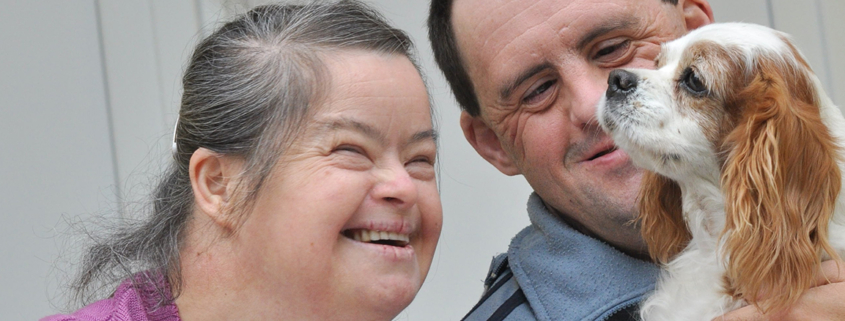 An older couple, both with Down syndrome, smile and look at a dog the man is holding