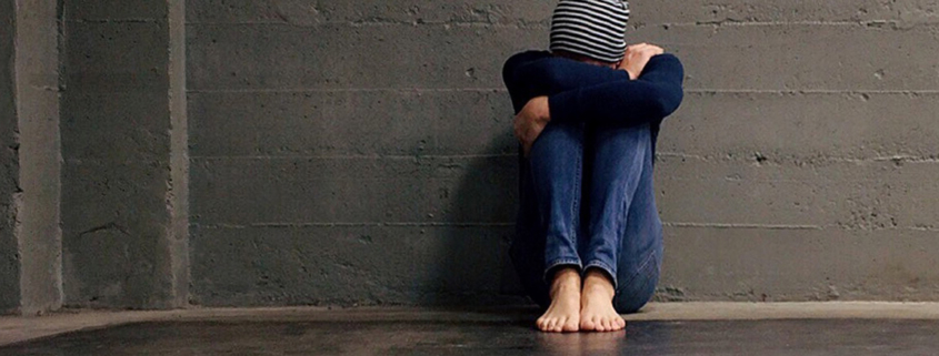 A distraught looking person sits on the floor with their head in their arms against their knees, and a brick and concrete wall behind them