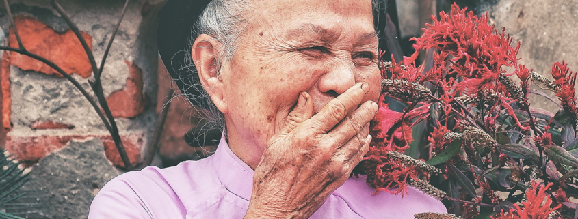 An elderly woman stands with her hand on her mouth in front of a brick wall with foliage and flowers beside her