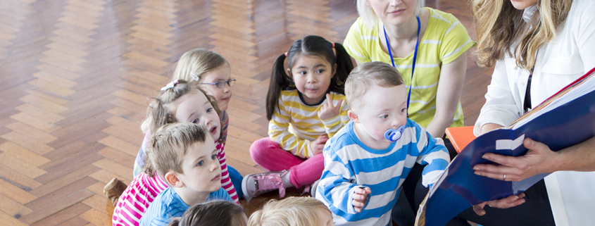 A small group of nursery school students sit around a teacher on a wood floor as she reads them a book