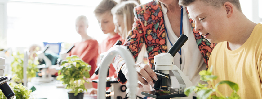 Middle-school aged students sit around a table at school with microscopes and plants. The teacher assists a boy slightly out of frame.