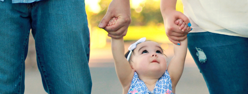 A baby holds the hands of both parents, looking up at them