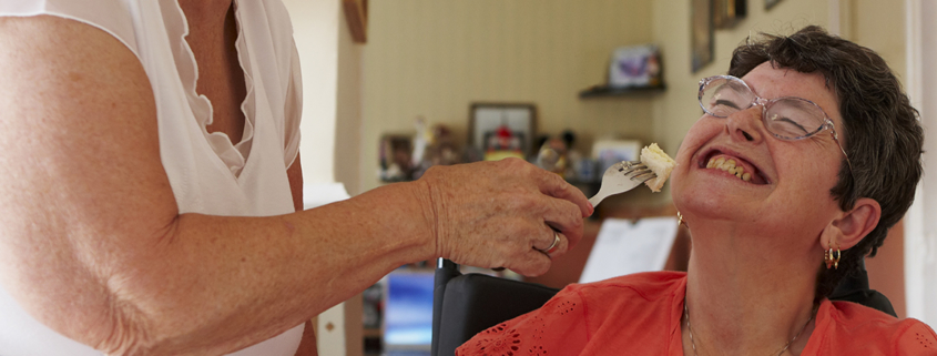 A caregiver holds a fork with food up to a seated woman who is smiling