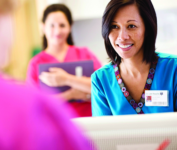 Two nurses, one in the foreground and one in the background, smile at a person who has their back turned to the camera