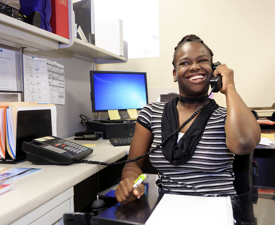 Young woman sits at desk in office and smiles while holding phone to ear