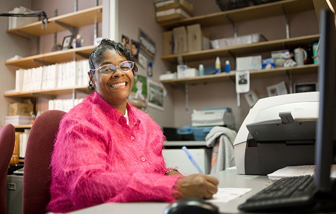 Older woman in glasses wearing a pink fuzzy shirt, sitting in front of a computer in an office setting