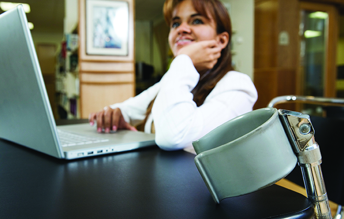 Smiling woman sitting in front of laptop in a library. Forearm crutch resting against table in the foreground.