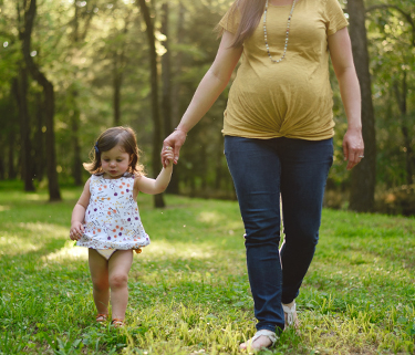 A pregnant woman walks through trees and grass, holding the hand of her young daughter