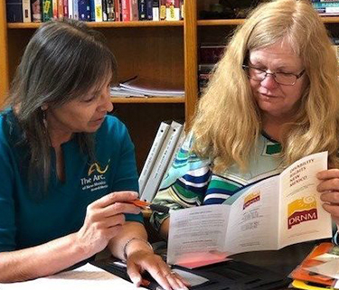 Two women sit at a table with a bookshelf in the background. One holds a pen and points to a brochure the other woman is holding open.