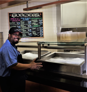 Kevin poses, smiling, in front of the food counter at work.