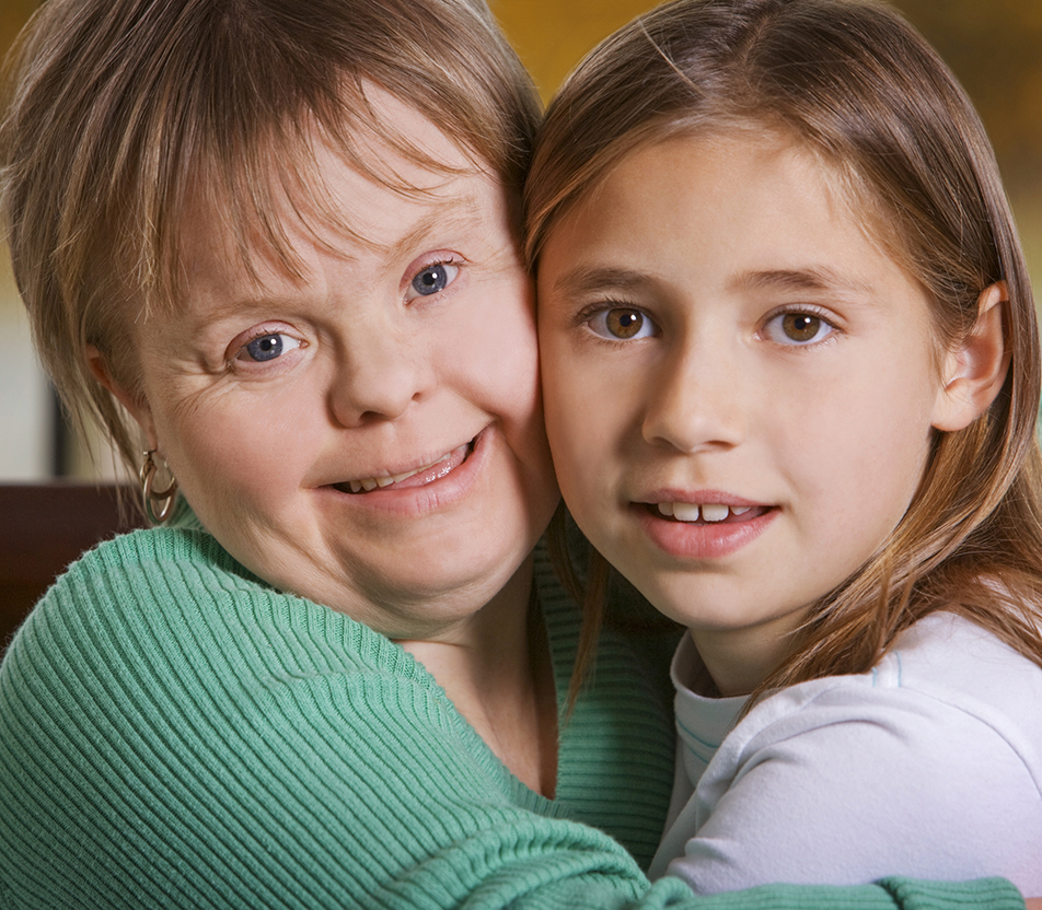 Two young women smile and pose for a close up shot. One is wearing a green ribbed sweater and the other is wearing a light purple shirt.