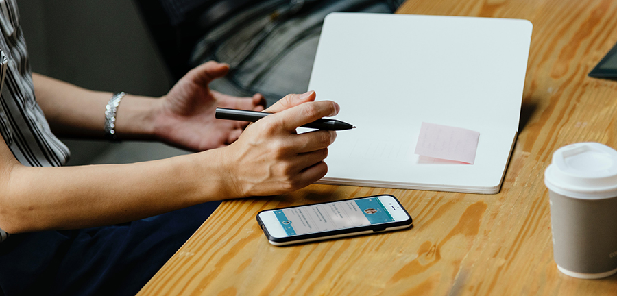 close up of a notebook on a table with a pink post it in it, with a coffee cup and phone nearby.