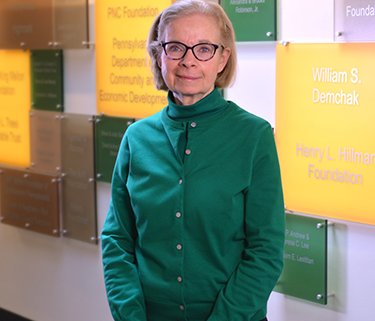 An older woman in glasses and a green turtleneck stands smiling at the camera with her hands clasped