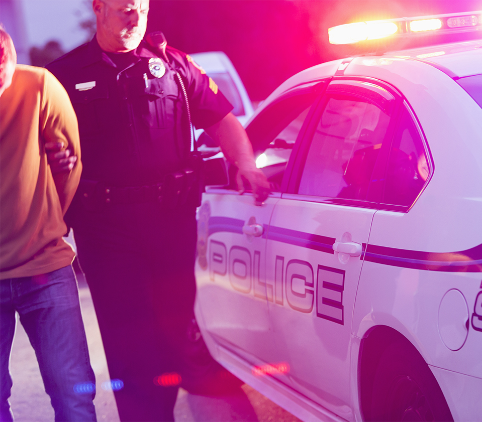 Police officer making an arrest, escorting a young man in handcuffs toward the back seat of his police car, at night.