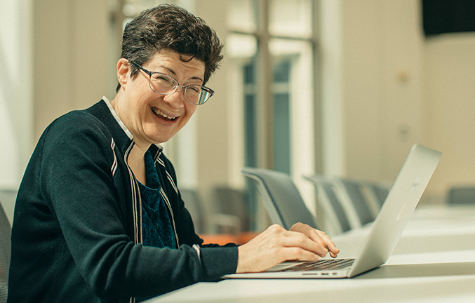 A woman sitting a conference room table on a computer turns to the camera and smiles as she types