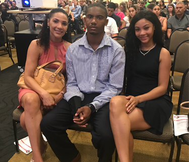 Two women sitting on either side of a man in the front of a conference room. They are all dressed up. There are many rows of people sitting behind them.