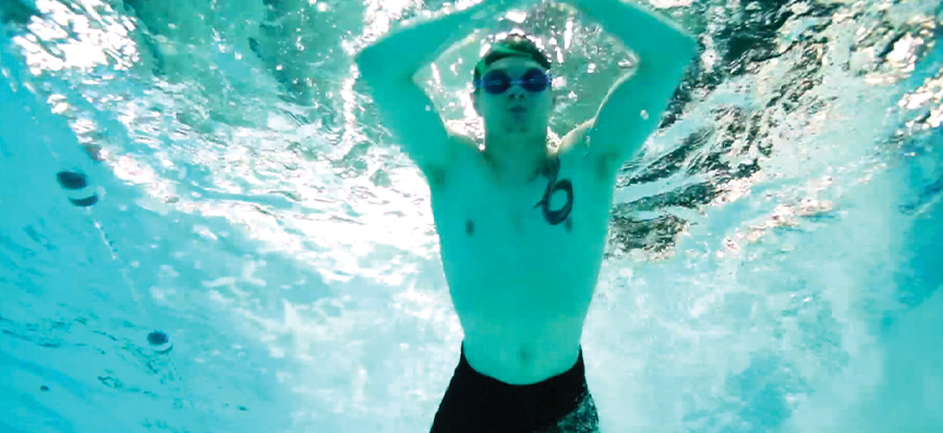 A man underwater in a swimming pool. He is wearing swim shorts, goggles and a swim cap and his hands above his head.