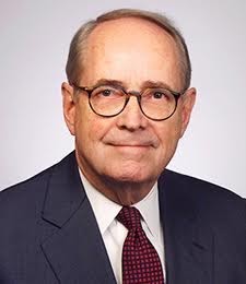 A headshot of an older man with glasses, wearing a suit and red tie. He smiles with his mouth closed. 