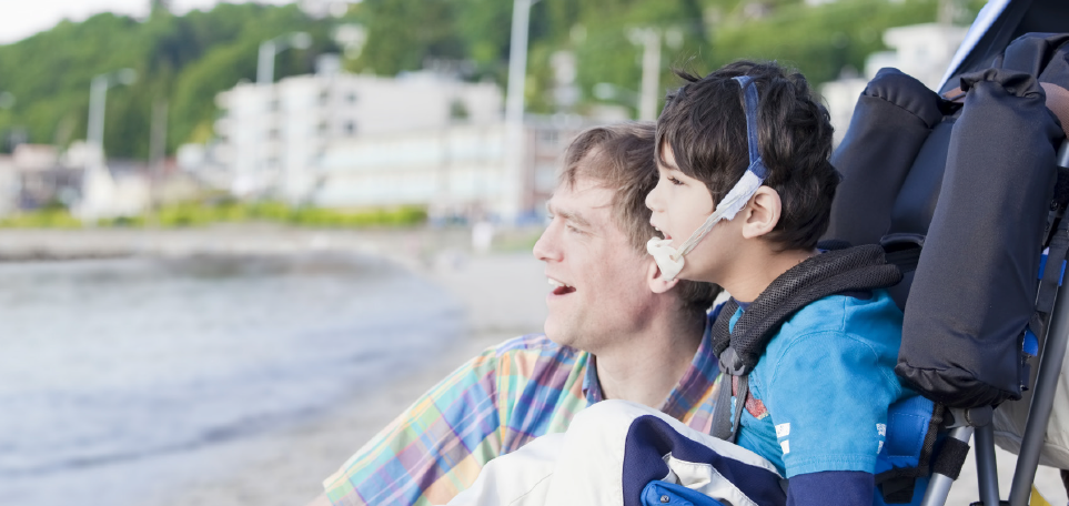 A man kneels next to his young son, who is in a wheel chair. They are at the beach, looking out at the water.