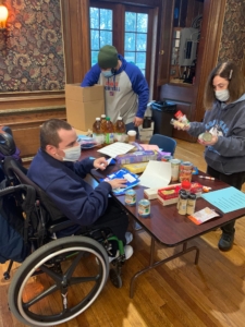 A volunteer in a wheelchairs sits at a table. He is working on meal kit assembly, with various items strewn across the table, wile other volunteers stand nearby helping. 