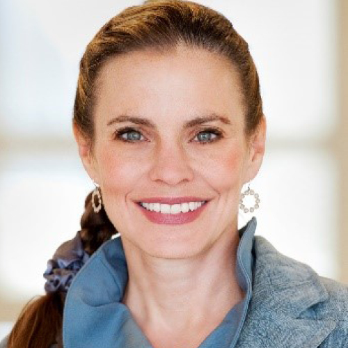 A woman with long brown hair in a side braid wearing a denim blazer witha blue blouse and earrings