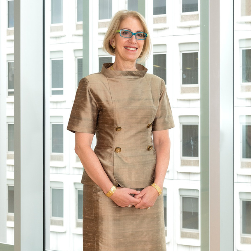 A woman with short blonde hair, glasses, and a gold dress stands smiling with her hands clasped in front of her