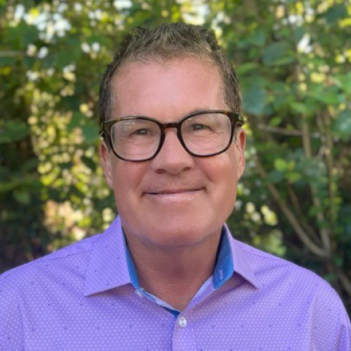 A man with short brown hair wearing a purple dress shirt and black glasses