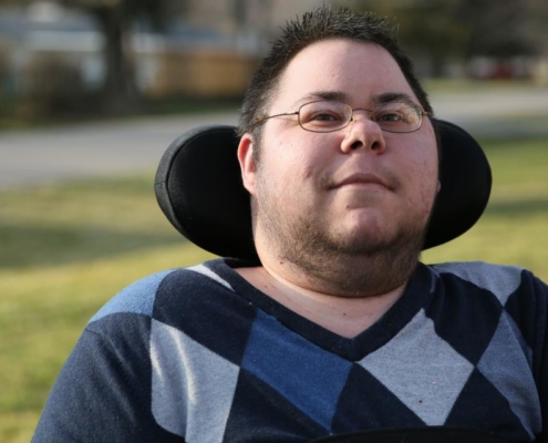 Dominick a masculine appearing person with green eyes and brown spiked short hair gives a small smile. He is wearing gold wire-rim glasses and has some stubble and hair along his cheek line. This headshot shows from his mid-chest area upward. He is wearing a blue, black, and gray Argyle long-sleeve shirt. He sits in his wheelchair, and his black headrest wraps around the back of his head. The background is blurred, but you can tell there is grass behind him and he is outside.