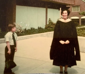 A woman in a graduation cap and gown stands on a paved area, with a young boy in a dress shirt and tie to her left looking at her.