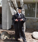 A graduate stands in front of a tree on a sandy spot with boulders around him. He is wearing a cap and gown, holding a diploma, and smiling.