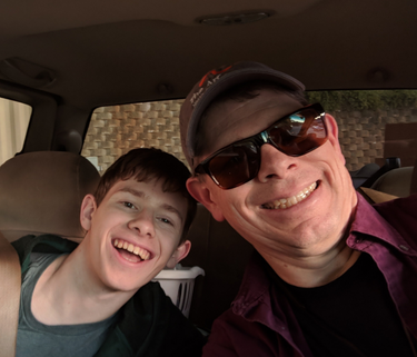 A selfie of a father and his teenage son, sitting in a car and smiling. The father is wearing sunglasses.