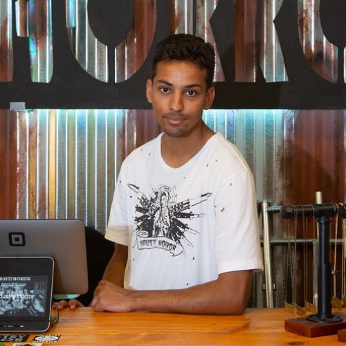 A young man with brown skin and facial hair in a white t shirt leans an arm on a wood counter and looks at the camera.