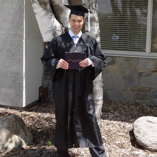 A man in a black graduation cap and gown stands holding his diploma and smiling. Behind him are some rocks, dirty, and a tree and a building with a window.