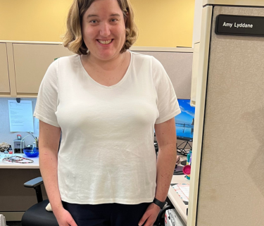 A white woman with blonde hair to her shouldersstands smiling with her arms at her sides in front of an office desk cubicle. She is wearing a white t shirt, black pants, and white shoes.