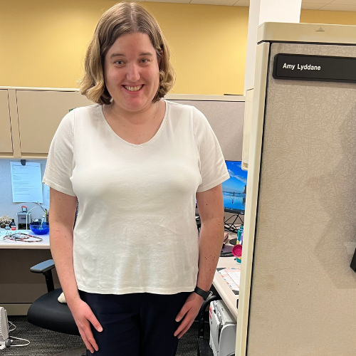 A white woman with blonde hair to her shouldersstands smiling with her arms at her sides in front of an office desk cubicle. She is wearing a white t shirt, black pants, and white shoes.