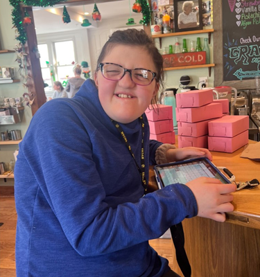A young adult woman with developmental disabilities is holding a tablet. She's looking at the camera and has a big smile.