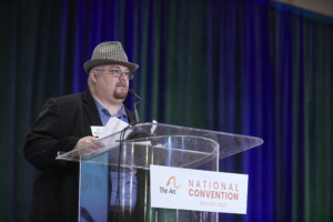 Mitch Routon stands a podium on a stage, speaking to an audience at The Arc National Convention.