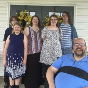 A man with disabilities is in a wheelchair. Next to him are his wife and young adult children. They are standing outside and smiling.